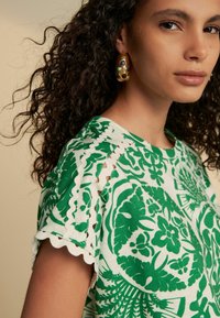 Green floral-patterned top made of soft fabric, featuring a scalloped neckline and sleeve edges with cut-out details. Complemented by bold earrings.