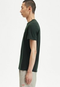 Camiseta de algodón de color verde oscuro con cuello redondo y mangas cortas. Presenta un pequeño logo en el pecho, textura suave y diseño ajustado.