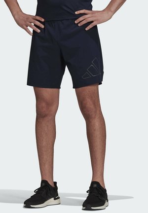 Mann trägt schwarze Sportshorts mit einem dezenten Logo am linken Bein und schwarze Laufschuhe, steht mit den Händen in der Hüfte.