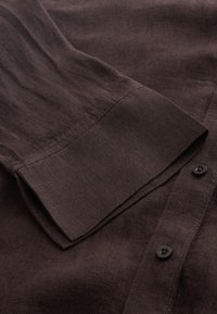 Manche de chemise marron foncé avec détail de poignet, boutons visibles et tissu texturé en gros plan.