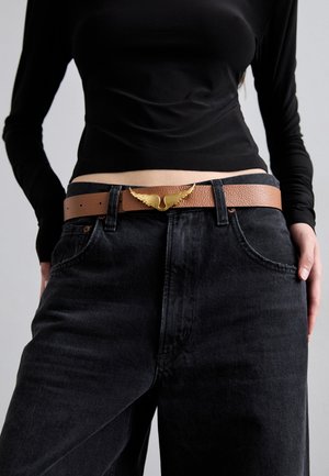 Brown leather belt with a textured finish, featuring a gold wing-shaped buckle, worn with black high-waisted jeans and a black top.
