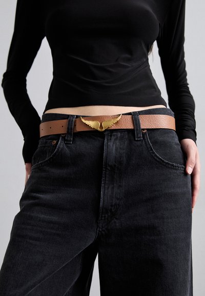 Cintura in pelle marrone con finitura testurizzata, dotata di una fibbia dorata a forma di ala, indossata con jeans neri a vita alta e una maglietta nera.