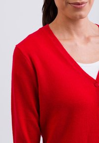 Röd ullkofta med V-hals, ribbade kanter och en enkel knappstängning. Slät textur med en figurnära design, delvis bärs över en vit topp.