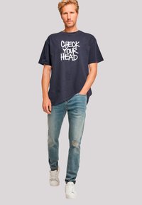 Navy graphic t-shirt with white "CHECK YOUR HEAD" text, paired with light blue distressed jeans and white sneakers. Casual fit design.