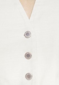 Vit skjorta i tyg med V-ringning, med tre silverfärgade tryckknappar prydda med "JEANS DKNV." Slät textur, minimalistisk design.