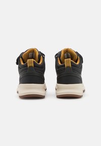 Pár černých a hořčicově žlutých sportovních bot s tlustými bílými podrážkami, pohled zezadu na čistě bílém pozadí.