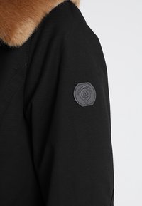 Czarna kurtka z futrzanym kołnierzem, wykonana z teksturowanej tkaniny, z okrągłą, szaro-skalowaną łatą, na której widnieje napis "Marc O'Polo" oraz "1967".