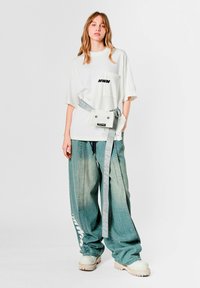 Camiseta blanca de gran tamaño con texto negro "MWM", combinada con pantalones anchos de mezclilla azul claro y un bolso cinturón gris con diseño de ojo y logo.