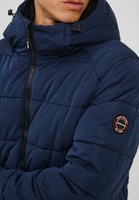 Marineblauer Puffer-Jacke mit hohem Kragen, ausgestattet mit einem Reißverschluss vorne, gesteppten Paneelen und einem runden Logo-Patch auf dem linken Ärmel.