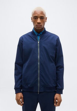 Mann mit kurzen blonden Haaren trägt eine dunkle, mit Reißverschluss versehene Jacke über einem blau gemusterten Hemd mit Kragen, steht vor einem einfarbigen, hellen Hintergrund.