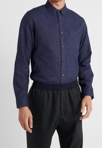 Man in een donkerblauwe overhemd met lange mouwen en knopen, ingetakt in zwarte elastische broek, staand tegen een effen lichte achtergrond.