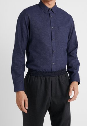 Homme portant une chemise à manches longues bleu foncé boutonnée, rentrée dans un pantalon noir à taille élastique, se tenant contre un fond clair uni.
