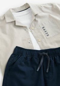 Weißes T-Shirt unter einem gestrickten beigen Hemd mit Kragen und Brusttasche, kombiniert mit navyblauen Shorts aus elastischem Material mit Kordelzug.