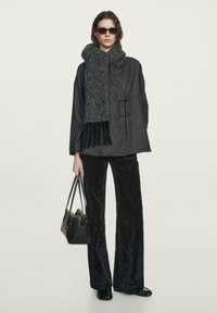 Sort uldblandingsfrakke med brede ærmer og stanglukninger, sammen med et langt, struktureret grønt tørklæde og skinnende sorte fløjlshyper.