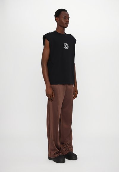 T-shirt preta sem mangas com um logótipo gráfico redondo, combinada com calças castanhas largas e sapatilhas pretas, contra um fundo neutro.