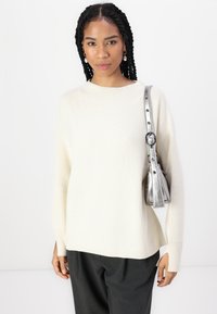 Pull en maille crème avec des manches longues, associé à un pantalon gris. Un sac métallique argenté avec une bandoulière est également présenté.