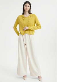 Gelber Cardigan mit rundem Kragen und Knopfdetails, kombiniert mit weiten cremefarbenen Hosen und beigen Absätzen, vor einem schlichten Hintergrund.