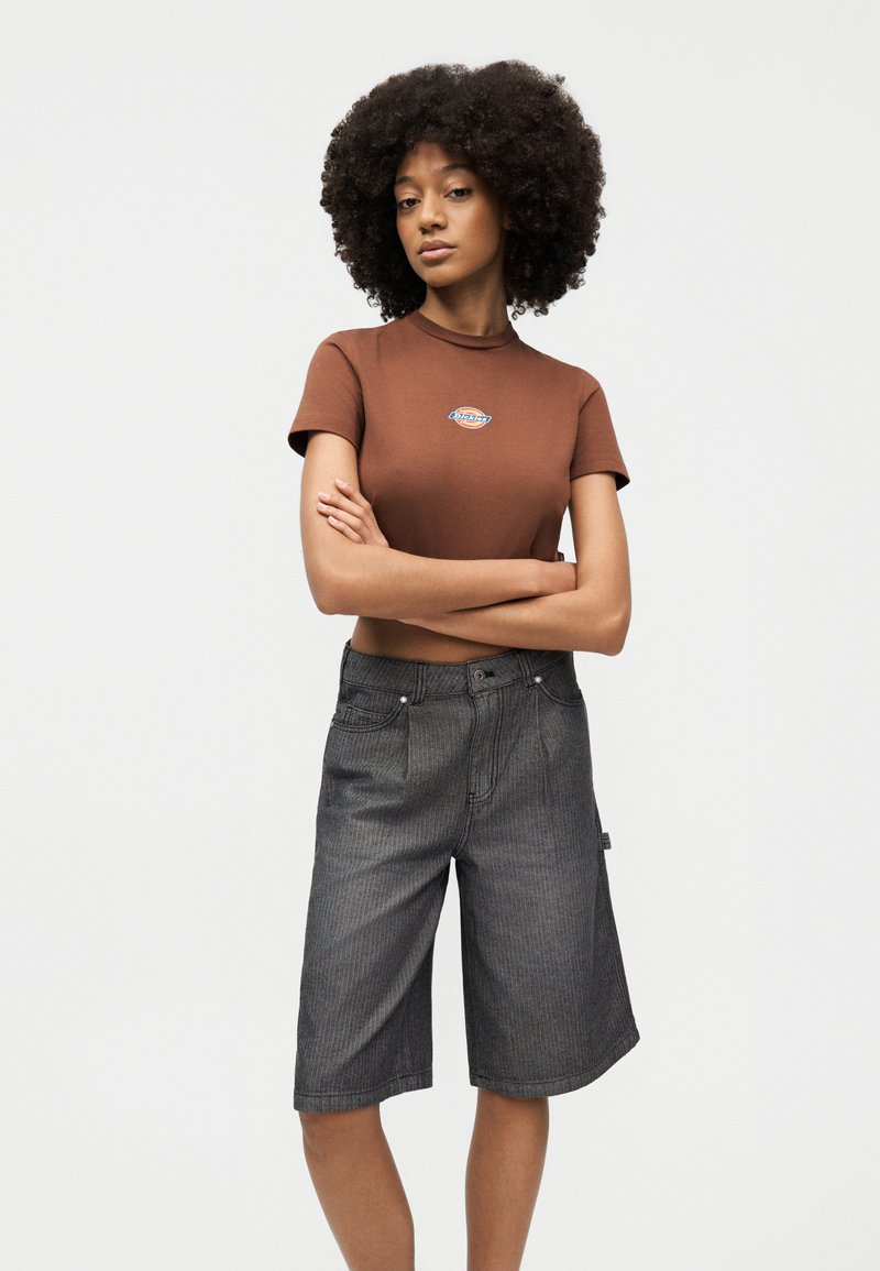 Giovane donna con capelli ricci che indossa un top corto marrone a maniche corte e pantaloncini grigi fino al ginocchio, in piedi con le braccia incrociate.