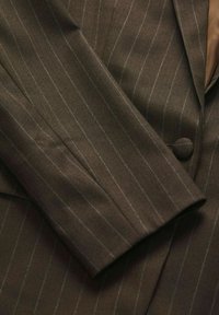 Rjav herringbone blazer material z tankimi vertikalnimi črtami. Ima gladko teksturo in gumb na sprednji strani.