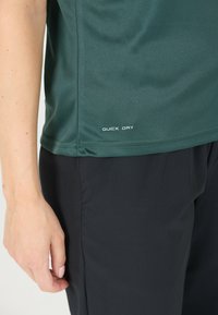 Mörkgrön snabbtorkande träningsskjorta med korta ärmar, med en slät textur och en tryckt etikett vid sömmen. I kombination med svarta byxor.