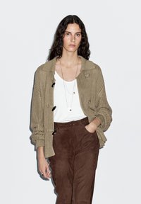 Beige stickad cardigan med krage och fyra svarta knappar, över en vit topp, kombinerad med byxor i brunt mocka. Enkel kedja inkluderad.