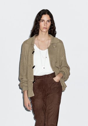 Beige gestrickte Strickjacke mit Kragen und vier schwarzen Knöpfen, über einem weißen Oberteil, kombiniert mit braunen Wildlederhosen. Einfache Kettenhalskette inklusive.