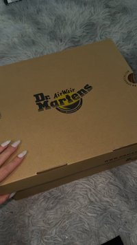 Cutie maro din carton pentru încălțăminte cu logo negru imprimat "Dr. Martens" și site web. Cutia are un mâner și este așezată pe o suprafață texturată gri.