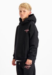 Zwarte katoenen hoodie met een frontzak, geribbelde manchetten en een rood logo op de borst. Model draagt het met bijpassende zwarte broek.