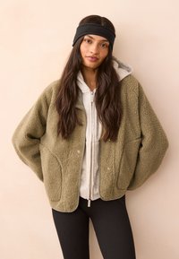 Maslinasto zelena jakna od flisa s teksturiranim završetkom, s kopčama na pritisak, nošena preko sive majice s kapuljačom na patentni zatvarač. Crna traka za glavu.