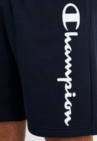 Marina idrottsshorts i texturerad tyg, med en stor vit "Champion"-logotyp vertikalt längs ena sidan och synliga sömdetaljer.