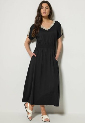 Kobieta stojąca z rękami w kieszeniach, ubraną w czarną sukienkę midi z beżowym wykończeniem w kształcie litery V oraz białe sandały.