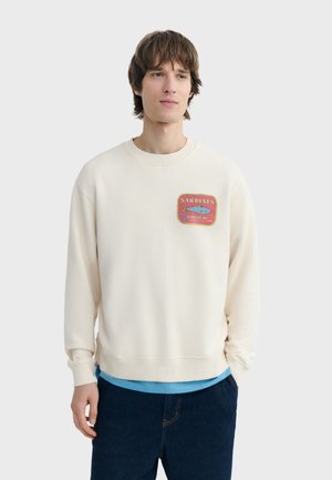 Młody mężczyzna ubrany w kremową bluzę z grafiką czerwonej puszki sardynek na piersi, nałożoną na jasnoniebieską koszulę i ciemne dżinsy.