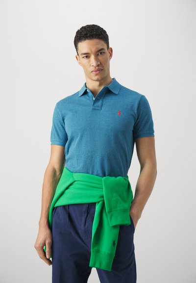 Camisa polo azul con cuello clásico, logo rojo y mangas cortas. Suéter verde atado a la cintura. El modelo lleva pantalones azul marino.