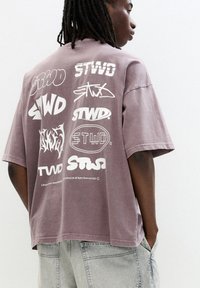 Overdimensioneret mauve t-shirt med forskellige hvide grafiske logoer trykt på ryggen. Stoffet ser blødt ud med en afslappet pasform og rund halsudskæring.