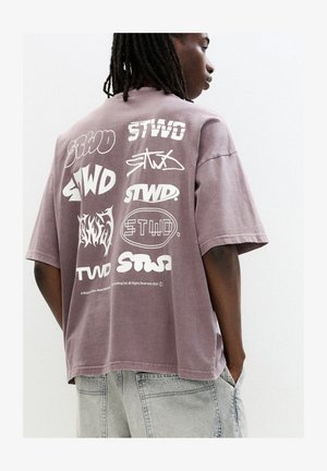 Overgrote mauve t-shirt met verschillende witte grafische logo's op de achterkant. De stof lijkt zacht met een relaxte pasvorm en ronde halslijn.