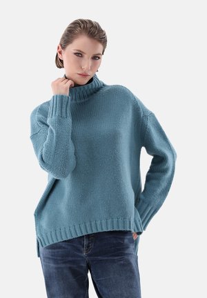Türkiser gestrickter Pullover mit hohem Kragen, gerippten Bündchen und seitlichen Schlitzen. Kombiniert mit dunklen Jeans, die einen lässigen Schnitt und Struktur zeigen.