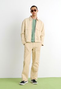 Beige bomullsjakke med to frontlommer, sammen med matchende beige bukser. Grønn t-skjorte under, svarte joggesko og solbriller.