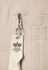 Beidzs tekstils ar vertikālu rāvējslēdzēju, tajā ir auduma birka ar uzrakstu "ALPHA INDUSTRIES INC. U.S.A." un teksturēta dūriena detaļa uz kabatas.