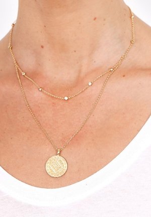 Deux colliers en or superposés autour du cou, l'un avec de petites pierres rondes et l'autre avec un pendentif circulaire texturé.