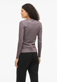 Top de manga larga en tejido metálico de color púrpura oscuro con un diseño ajustado, textura suave, cuello redondo y un brillo sutil. Combinado con pantalones negros.