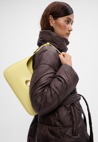 Smeđi pufasti kaput s visokim ovratnikom; žuta torba za rame zaobljenog oblika s zlatnim detaljima. Glatka tekstura; minimalistički dizajn.