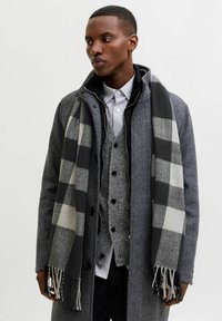Abrigo de lana gris sobre un chaleco gris, camisa a rayas y pantalones negros. Una bufanda de cuadros en blanco y negro cae sobre el abrigo, con bordes desgastados.