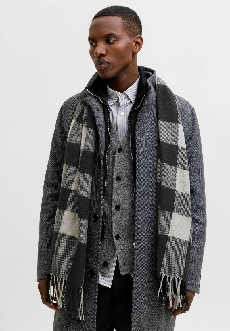 Abrigo de lana gris sobre un chaleco gris, camisa a rayas y pantalones negros. Una bufanda de cuadros en blanco y negro cae sobre el abrigo, con bordes desgastados.