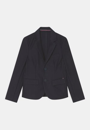 Blazer azul marino entallado con solapas de muesca, dos bolsillos delanteros con solapa, dos botones y una pequeña etiqueta roja, blanca y azul en el borde del bolsillo.