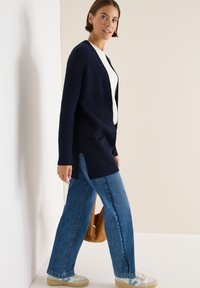 Marinblå ribbad cardigan med sidofickor, bärs över en vit topp. Matchas med ljusblå vida jeans och beige sneakers. Brun axelväska.