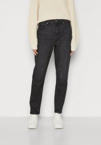 Femme portant un jean gris foncé taille haute, un pull en maille crème et des baskets blanches, debout devant un fond clair uni.