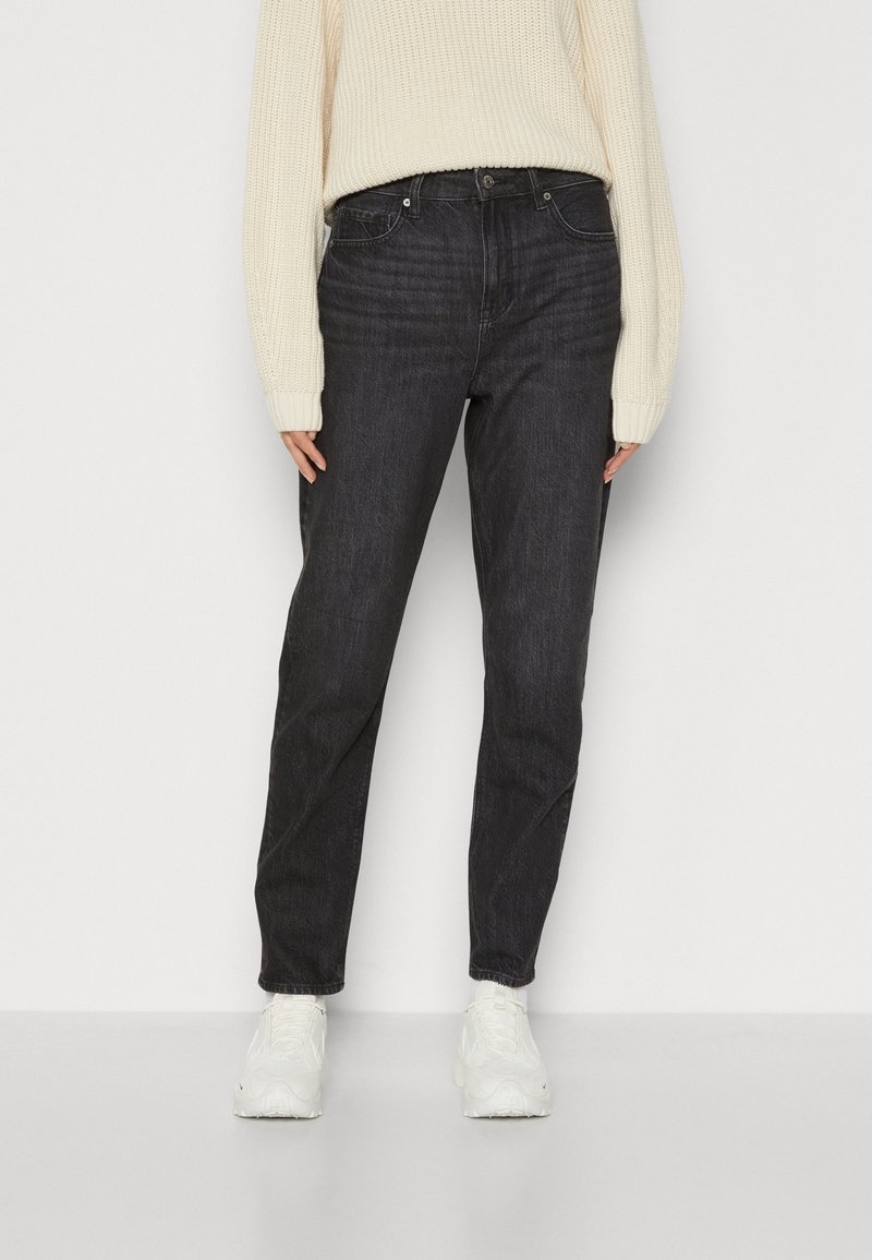 Femme portant un jean gris foncé taille haute, un pull en maille crème et des baskets blanches, debout devant un fond clair uni.