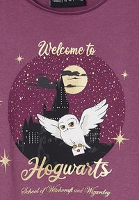 Purple T-shirt featuring a graphic of a castle, an owl holding a letter, and text. Gold accents and star details enhance the design.