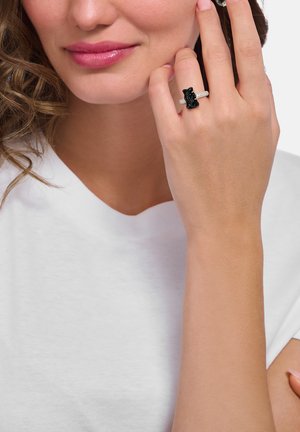 Black bear-shaped ring with a textured band, featuring a dark gemstone in the center. Model wearing a white shirt, hand resting near the face.