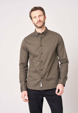 Man met baard die een bruin patroon-knopenoverhemd en zwarte broek draagt, staand tegen een effen lichte achtergrond.